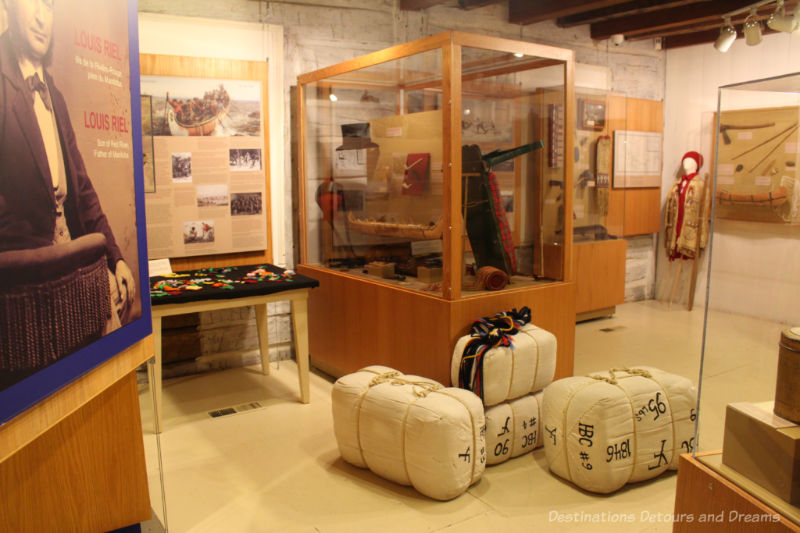 Room with displays (canoe models, clothing, packed parcels, written information) showcasing voyageur life and history at Saint Boniface Museum