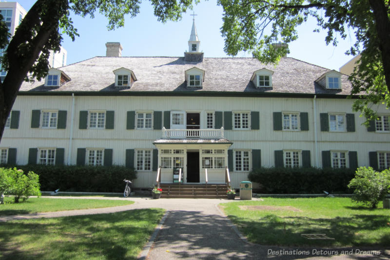 Two story white oak log building with green window shutters is a former convent housing St Boniface Museum showcasing French-Canadian and Métis heritage