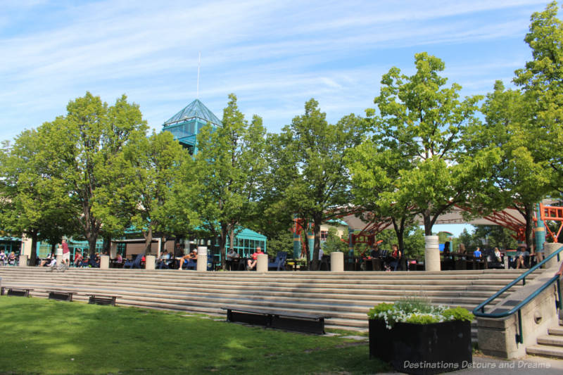 Grassy area in front of rows of stone steps/seating and upper level patio area at The Forks in Winnipeg, Manitops