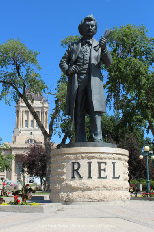 Full length statue of Louis Riel looking very statesmanlike on top a Tyndall stone base with the word RIEL on it.