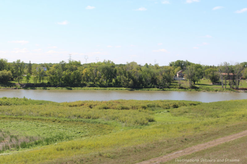 Calm, muddy river running through prairie with trees and houses on one side of the river