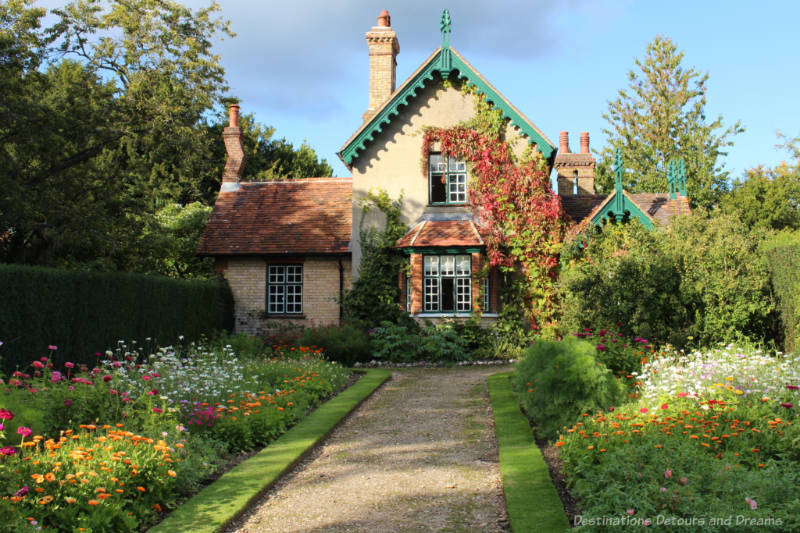 Gravelled pathway lined with massed of blooming flowers leading to a gingerbread house style stone cottage 