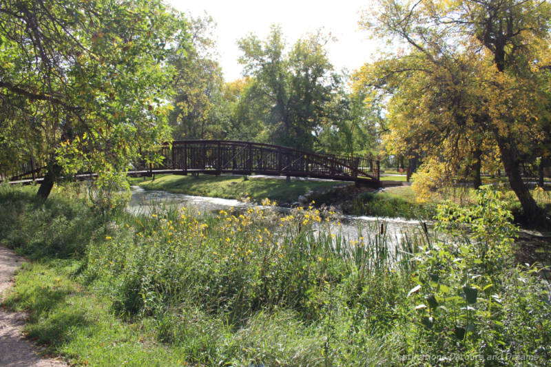 Brown foot bridge crossing a small river into a grassy treed park