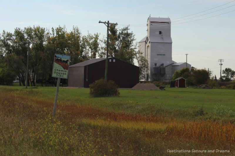 White Dufresne grain elevator behind a field with the Dawson Trail sign in it