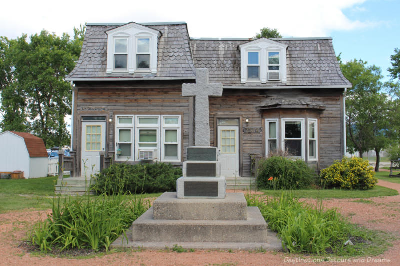 Stone cross memorial in front of old wooden building at Place St. Norbert in Winnipeg