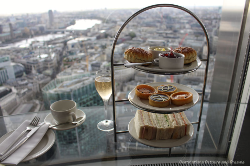 Cup of team, flute of champagne, and tiered tray of afternoon tea treats on a glass bar in front of window overlooking London at Vertigo 42