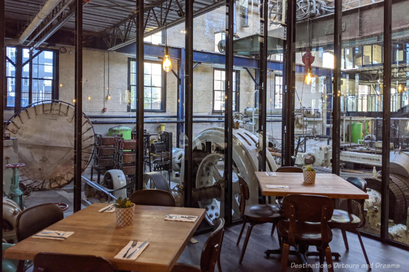 Tables inside a restaurant with century-old pumping station equipment on the other side of glass walls behind the tables at James Avenue Pumphouse Food and Drink