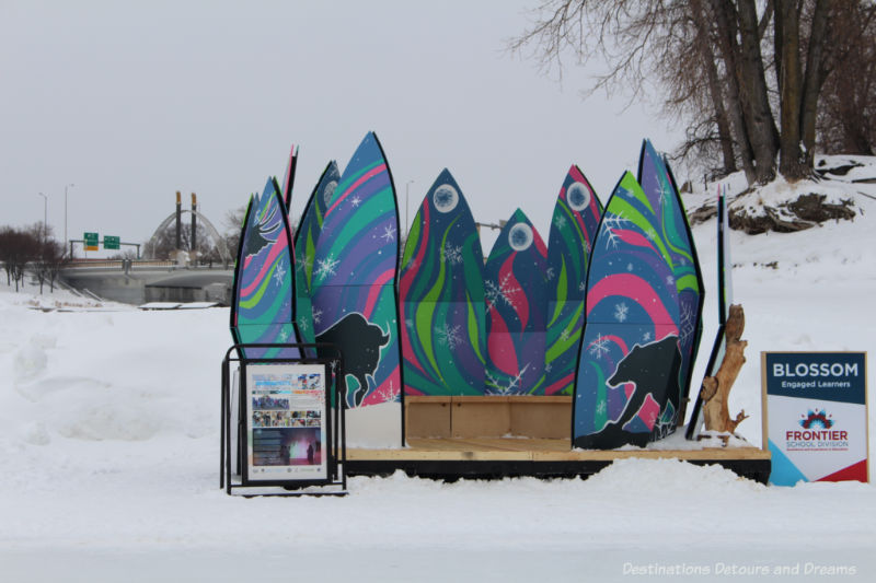 Colourful pointed panels decorated with stylized northern lights and northern wildlife form a warming hut on the river