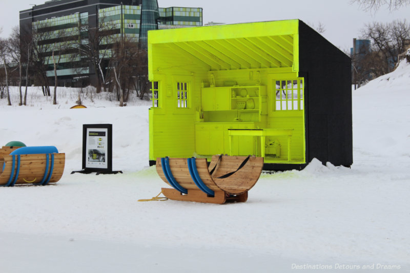A neon yellow three-walled warming hut designed to resemble the kitchen of a wilderness cabin