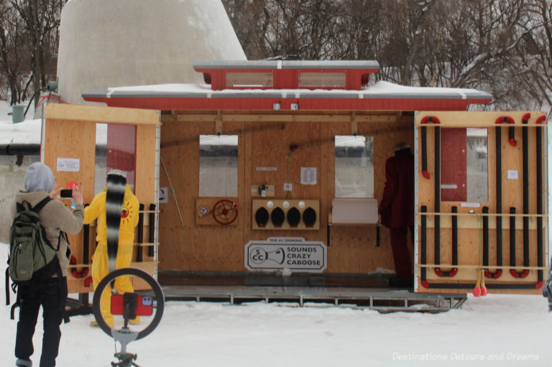 Caboose-like warming hut with one side wall opened up to reveal performers playing music-making gadgets