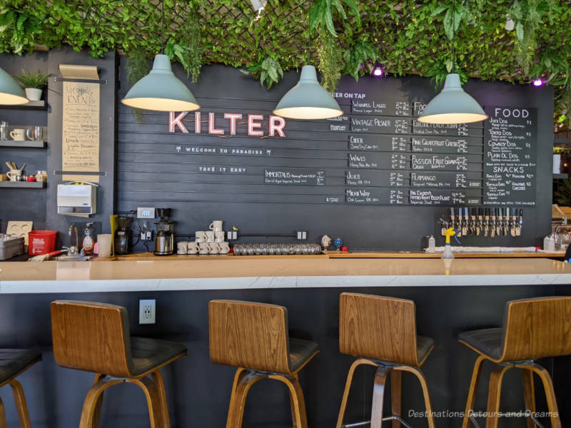 Bar at Kilter Brewing, a Winnipeg craft brewery, with menu on chalkboard like panel behind bar topped with greenery