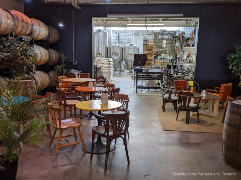 Round tables and chairs in the Winnipeg craft brewery Kilter Brewing taproom with wooden kegs of beer along one wall and an opening in the wall showing the brewery in the back