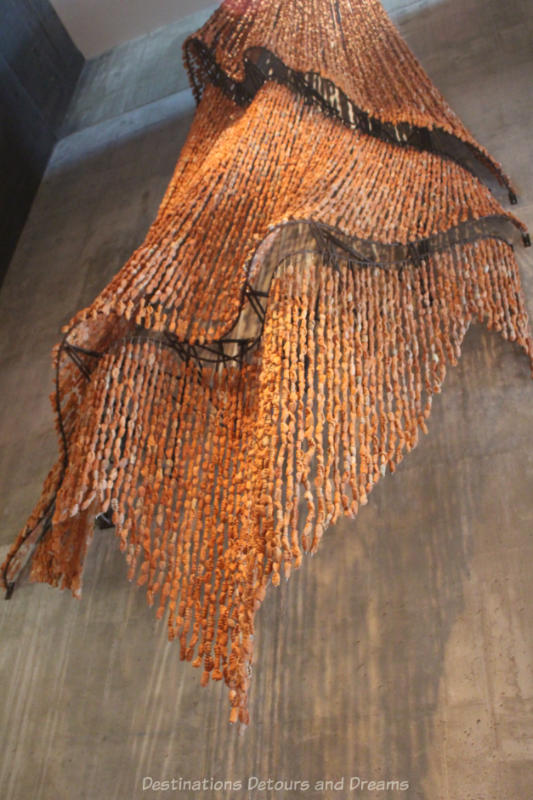 A hanging sculpture, Trace by Rebecca Belmore, made of clay bead strung together to look like a blanket hangs in the CMHR Indigenous Perspectives gallery