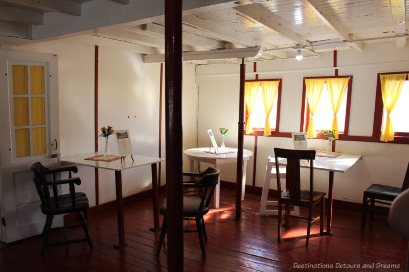 Tables with white tabletops and wooden chairs in a boat dining room with dark wooden walls, white walls, and square windows.