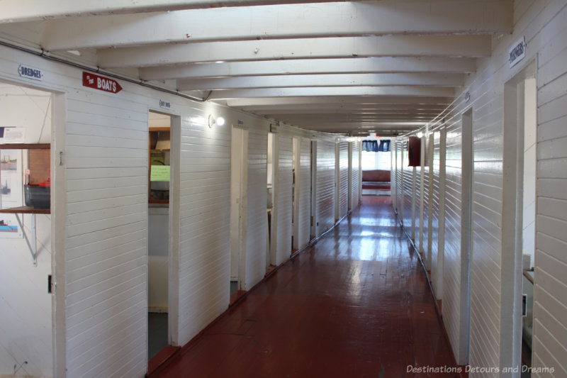 Ship hallway with dark wooden floors and wood white walls off which are the passenger cabins.