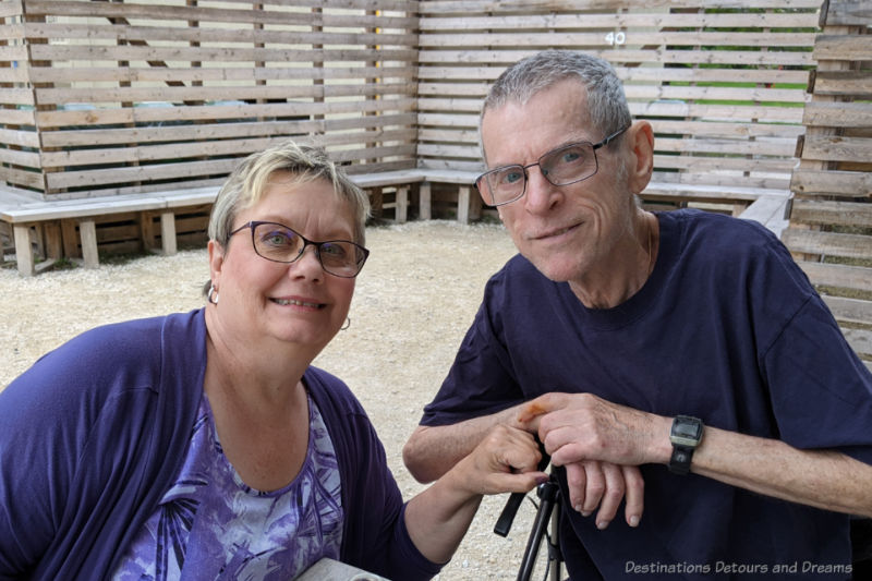 Man and woman smiling as they sit in a graveled patio area with wooden benching behind them