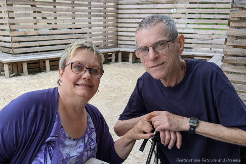 The Beer Can Enjoyment Man and woman smiling as they sit in a graveled patio area with wooden benching behind them