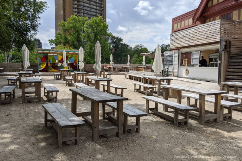 Graveled patio area with wooden tables and benches with food and drink counter to the side and mature green trees in the background