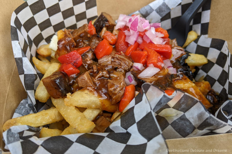 Poutine in a black and white checkered paper-lined contained - french fries covered with onions, red peppers, brisket, cheese and gravy
