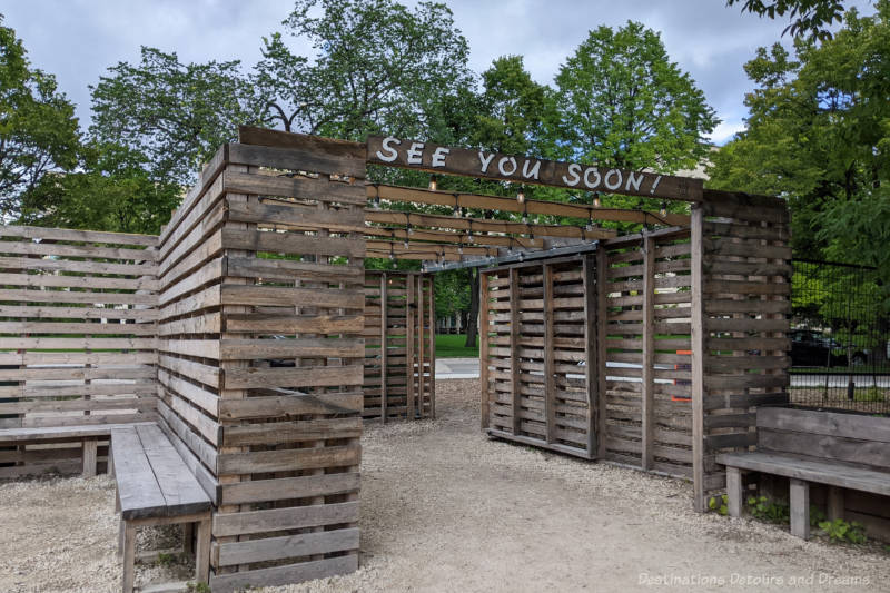 Wooden slat walls of an entrance/exit to a patio are with the letters See You Soon printed on top