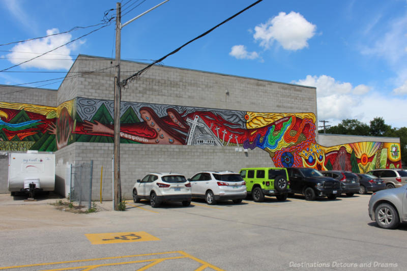 A brightly coloured murals with reds, oranges, yellows, and greens in panel panels spread across the long side of a building features residential school stories