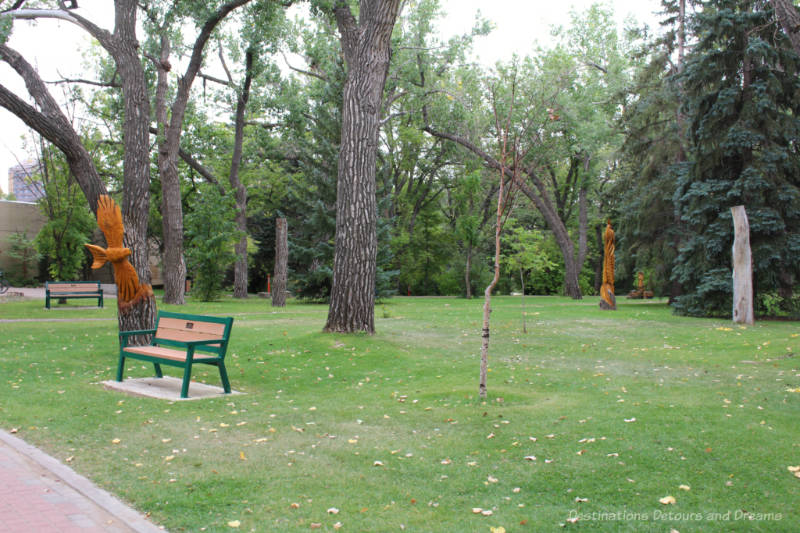 A park area with trees and benches and some art carvings on stumps of former trees