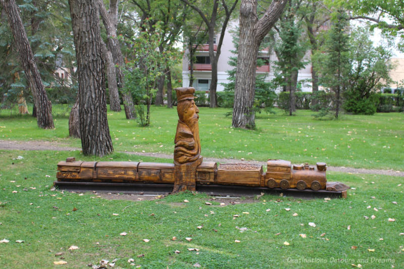 A wood carving in a park features a old man sitting atop a model of a train with six cars
