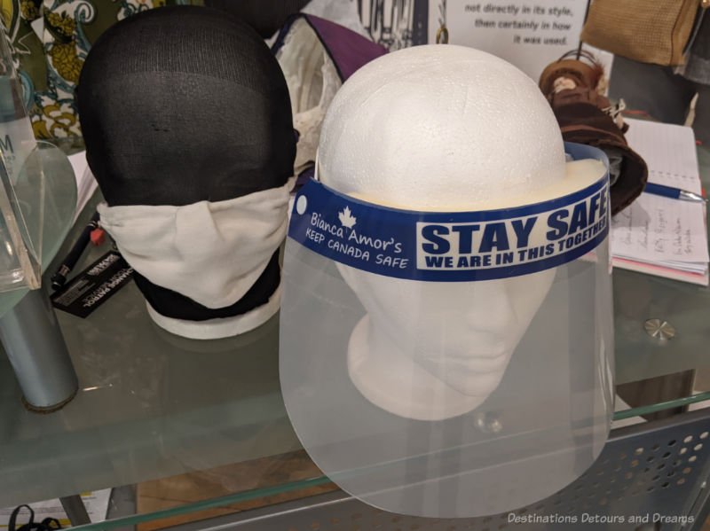 Mannequin heads sitting on a table and wearing PPE: a face shield on one, a white cloth face mask on the other.