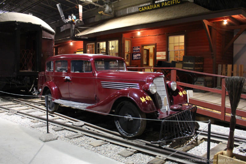 Museum display of old car equipped with rail wheels on a rail track in front of a rail station
