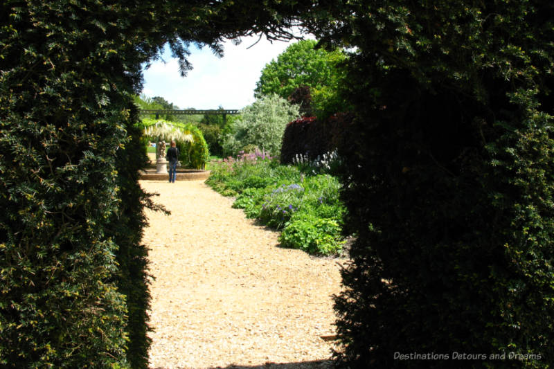 Looking through a greenery lined archway into a garden with a garden path down the centre bordered by flowering bushes and leading to a statue
