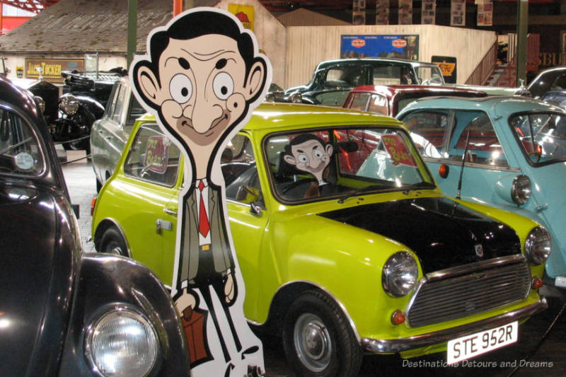 Motor museum exhibit of lime-yellow mini car used by Mr. Bean in the TV series with a cardboard cutout of Mr. Bean in the driver's seat