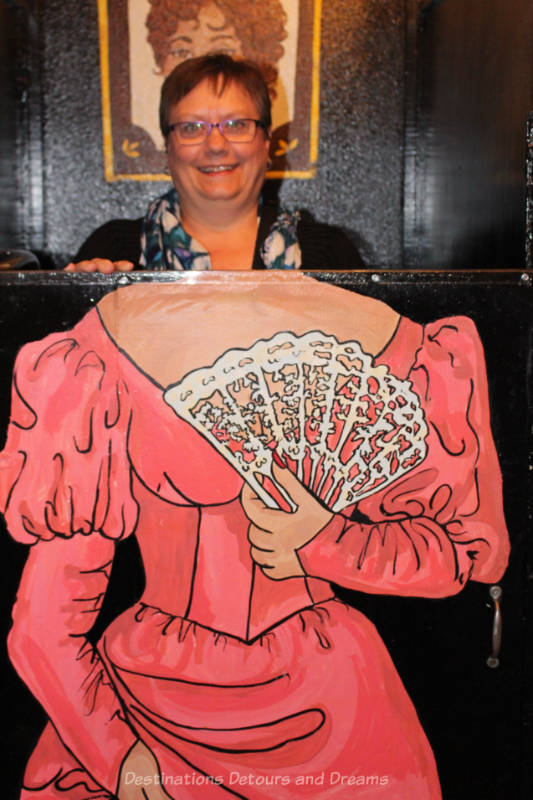 Figure of an Old West dance hall girl holding a deck of cards on a chest-high toilet stall door with the head of a present-day woman showing above the top of the body