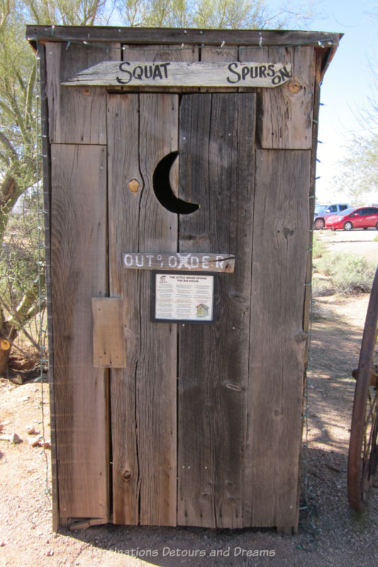 Wooden out house with a crescent moon peep house, an out of order sign, and signage on top of door that says Squat Spurs On