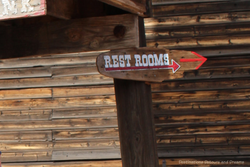 Wooden sign against an old wooden building with the rest rooms printed in white and a red arrow pointing right