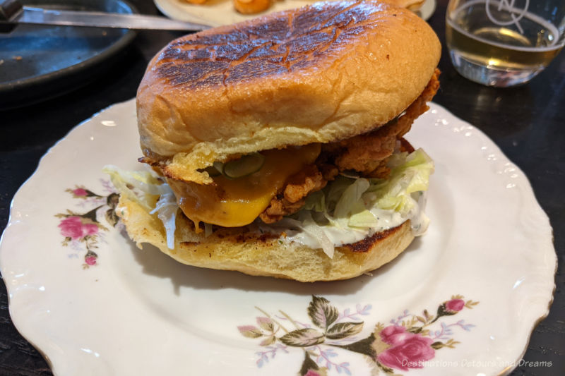 Fried chicken sandwich in a bun with lettuce, cheese, and pickles served on a pretty china plate with roses on it