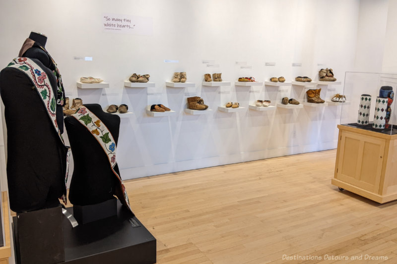 White wall with small white shelves displaying a collection of Indigenous beaded footwear