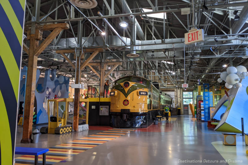 Train locomotive in centre of auditorium -like space of a children's museum with various colourful exhibits on either side