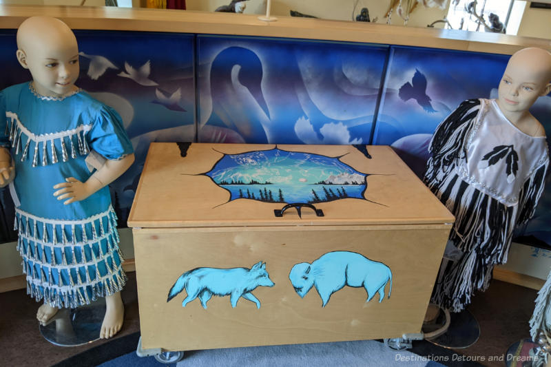 Chest decorated with Indigenous art and 2 child mannequins wearing beading clothing at Manitoba Indigenous Cultural Education Centre