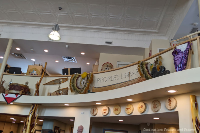 Curved wall with ledge below a mezzanine level with assorted Indigenous artifacts on display, such as canoe, snowshoes, poles, drums, and art