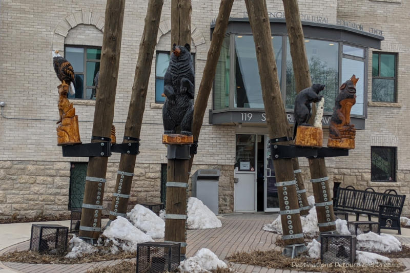Sculpture in front of Manitoba Indigenous Cultural Education Centre consists of seven large poles tilting inward in a circle with carved animals resting on shelve partway up the poles