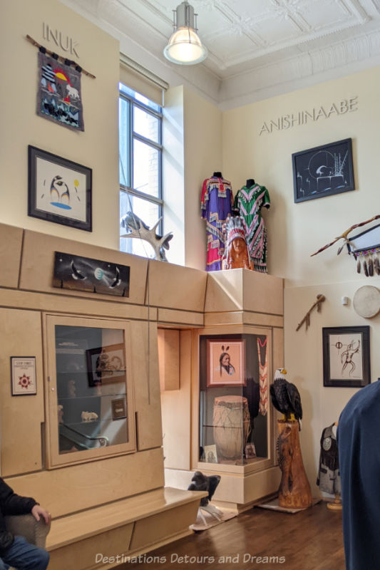 Indigenous art and artifacts on display on almond-coloured two-story high walls with shelve midway displaying more items