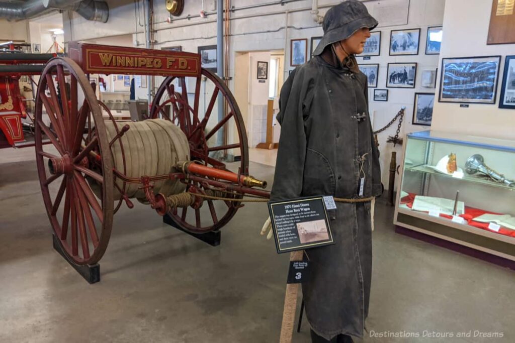 Mannequin in fire fighting apparel in front of 1870 hand-drawn hose-wheel wagon in museum display