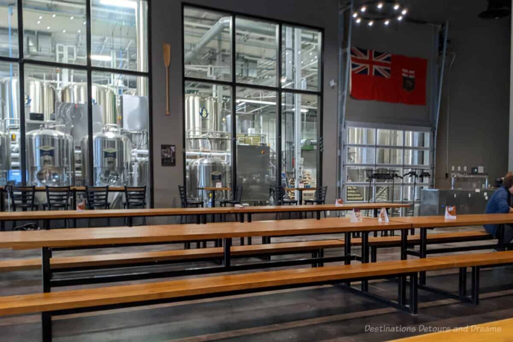 Interior of a tap room with picnic-style tables and a windowed wall providing views into the brewery