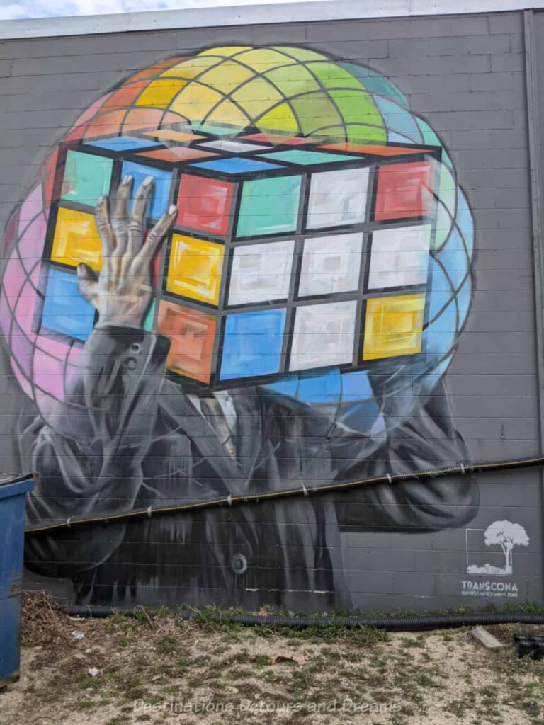 A side of building mural features the top half of a man in a suit holding a Rubik's Cube inside a multi-coloured orb than covers his head