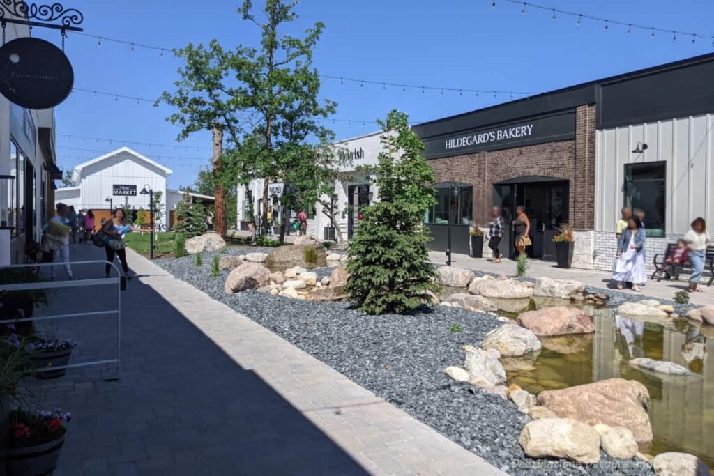 Outdoor pedestrian walkway between row of stores separated by landscaped boulevard at The Village at Pineridge Hollow