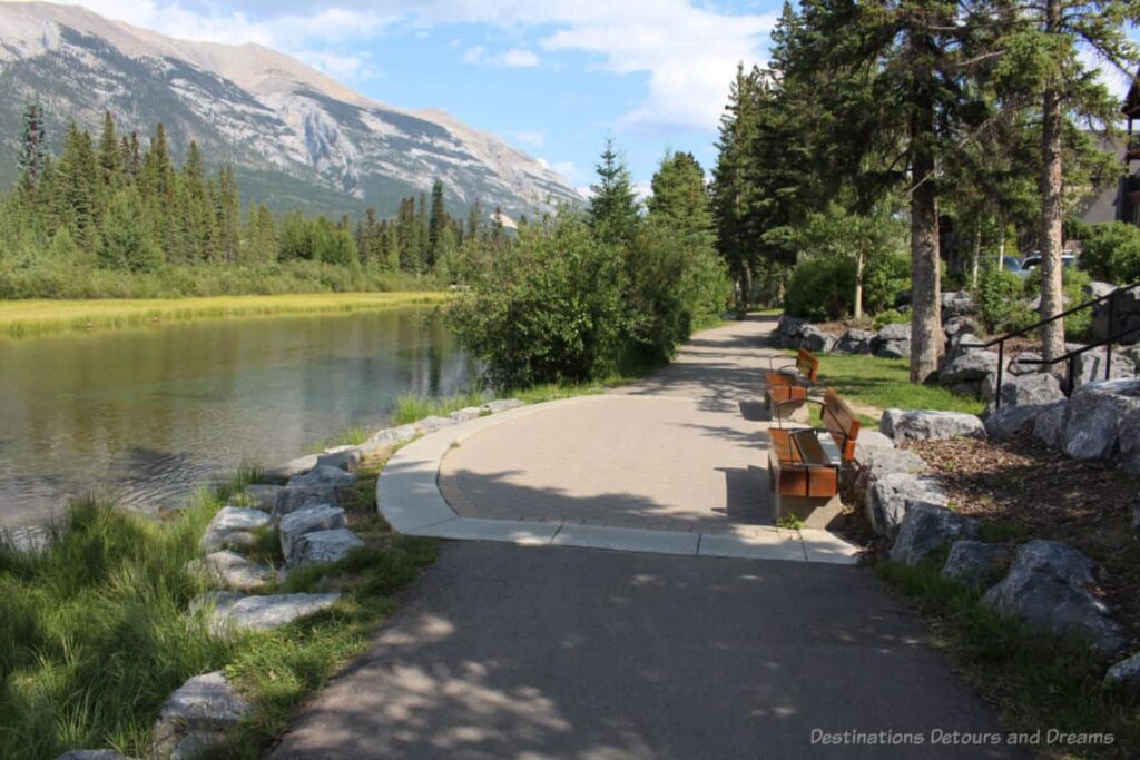 Creek lined with evergreens, view of mountains on one side, walking path with benches on the other