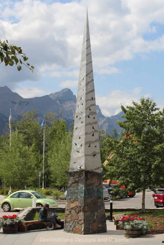 Tall pointed tower of a public art piece featuring painting of birds on the town and a stone design motif at bottom