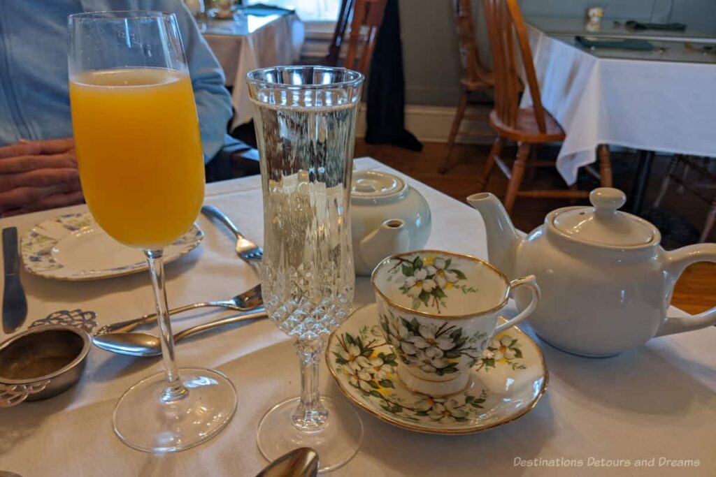 Table top containing white tea pots, a china cup and saucer, a glass of sparkling wine, and a glass of Mimosa