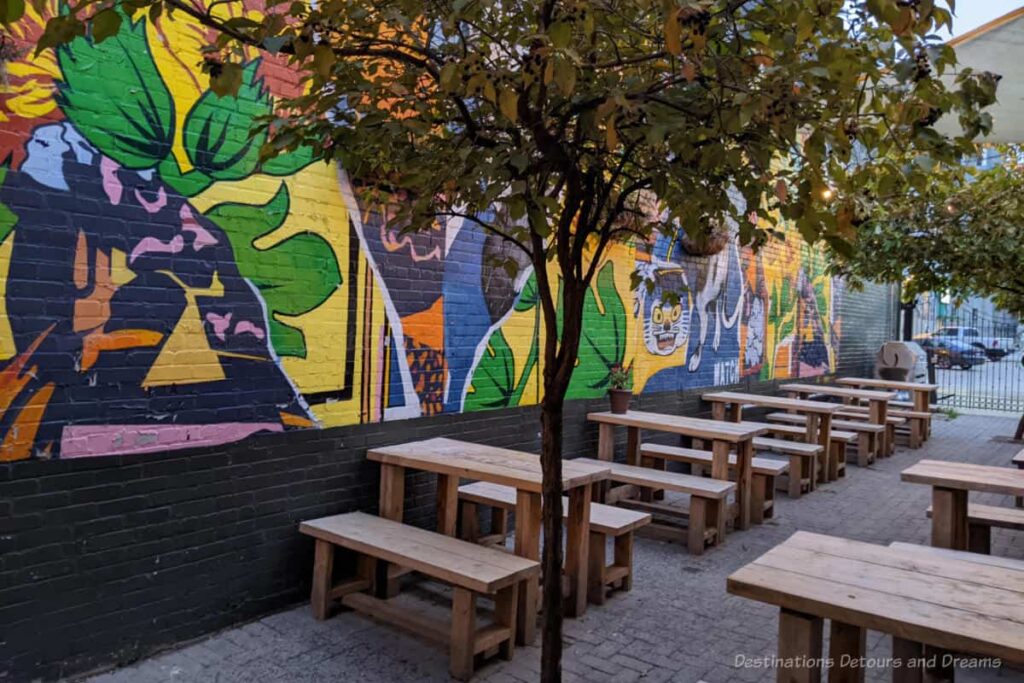 Side courtyard of a restaurant with wood tables and benches and a colourful mural on the brick wall of the neighbouring building