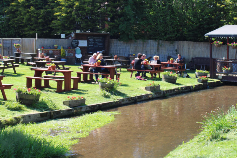 English pub garden area featuring picnic tables on grassy area beside stream 